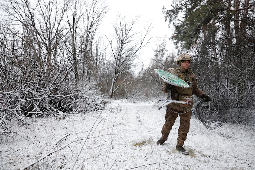 Man in military uniform carries satellite through snow.