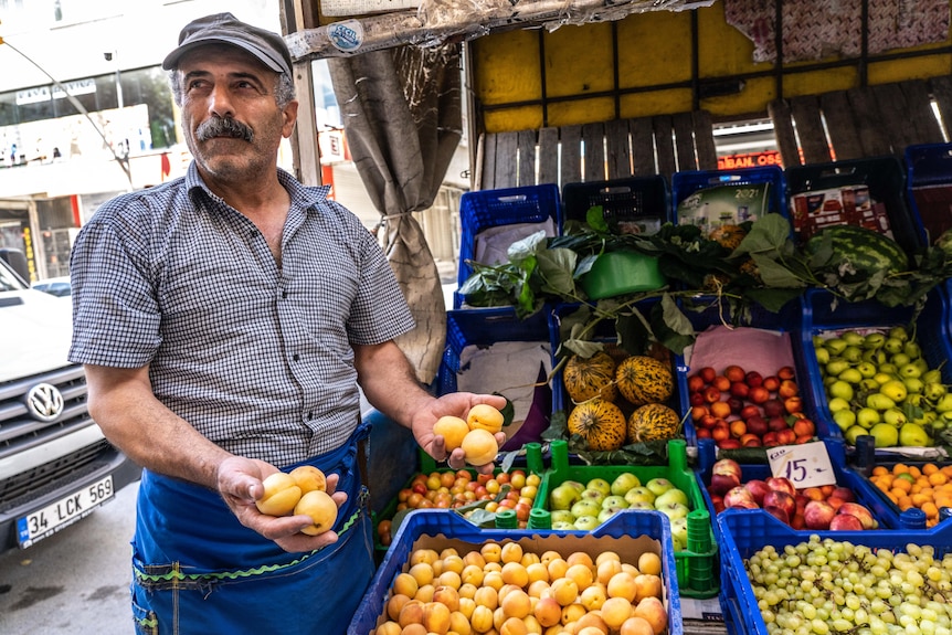 Un uomo con baffi folti con un cappello grigio e un grembiule blu tiene in mano delle albicocche, accanto a un chiosco di frutta