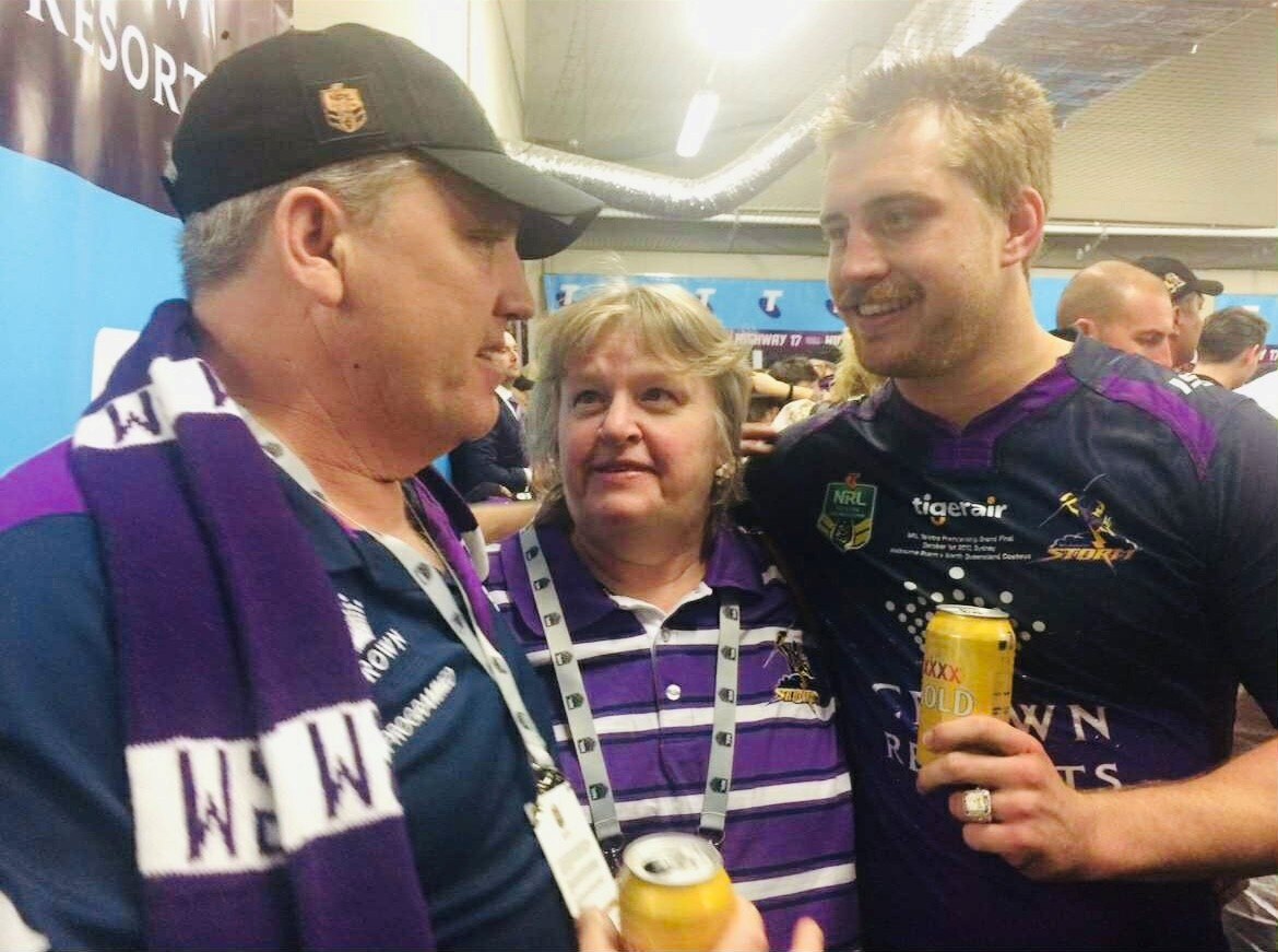 Smiling Steven, Debbie, and Cameron in purple and white football uniform embrace holding a beer, crowds behind him.