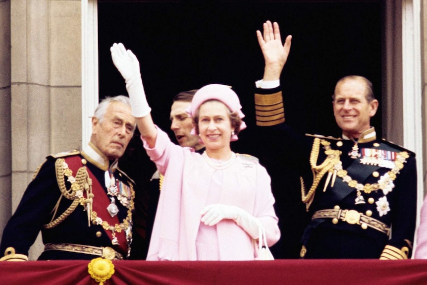 The Queen wearing a pink dress and hat, and white gloves, waving from the balcony beside Prince Philip.