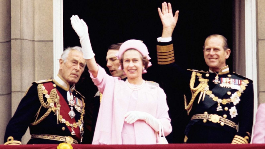 The Queen wearing a pink dress and hat, and white gloves, waving from the balcony beside Prince Philip.