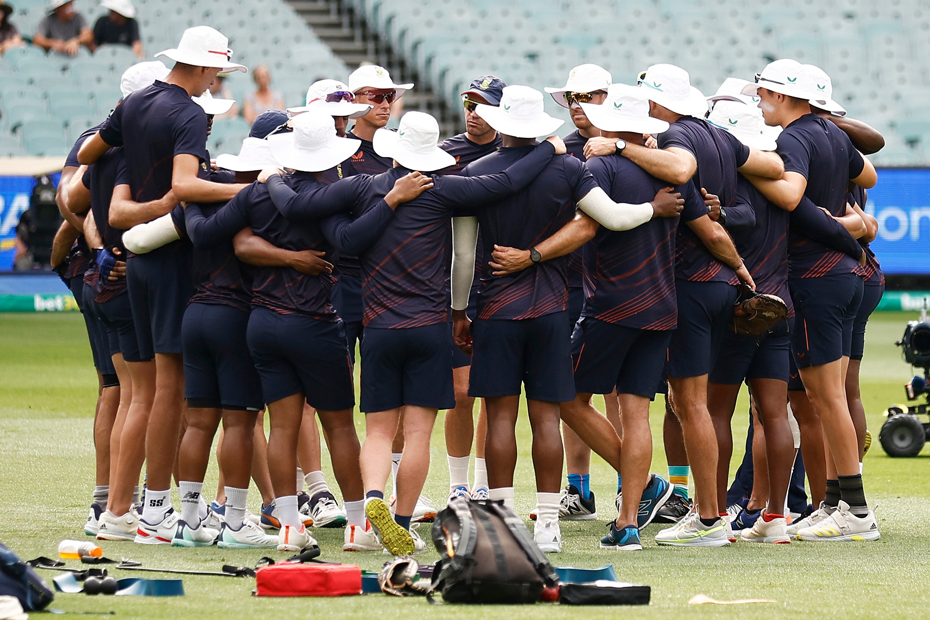 A group of cricket players wearing dark clothes and white hats gather in a circle