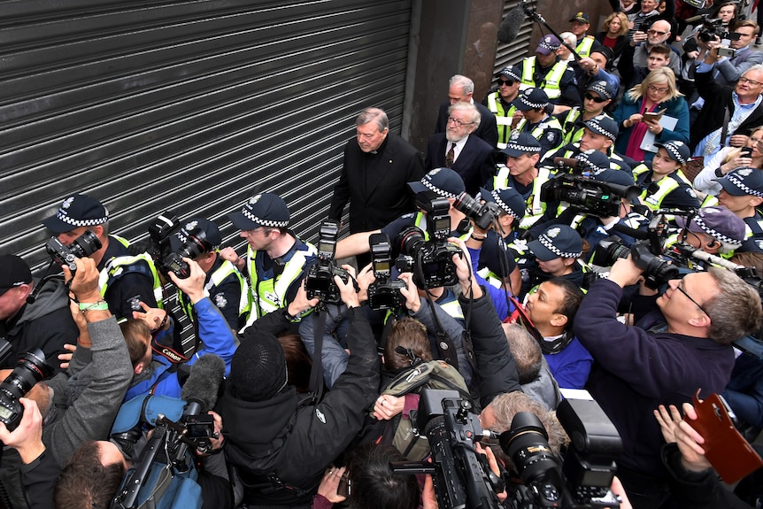 Man walking surrounded by press pack and police officers