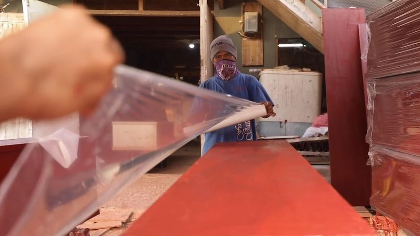 A coffin being covered in plastic by two men wearing full protective clothing. 