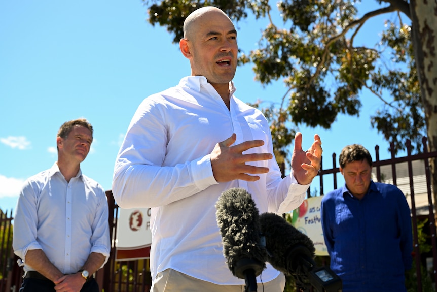 A bald man talking at press conference to journalists with other men standing around 