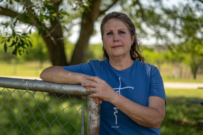 A white woman in a blue T-shirt leans on a fence post post outside