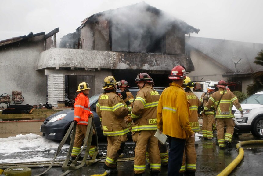 A group of firefighters stand outside a home destroyed by fire after a plane crashed into it.