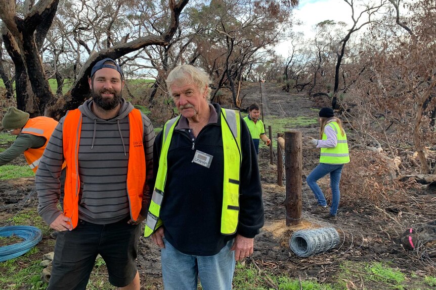 Blaze Aid volunteers Jack Tourlamain and Graham Norfolk stand in front of trees and wooden fence posts