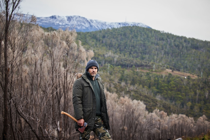 A man in his mid 40s in winter outdoor gear, including a beanie and jacket, holding an axe standing in front of forested hills