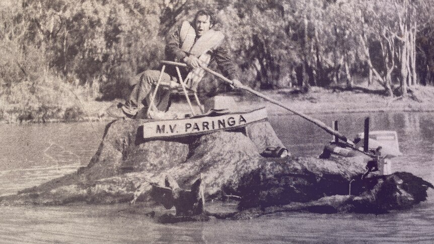 A man wearing a life jacket is on top of a tree stump with a motor attached, sailing on the river.