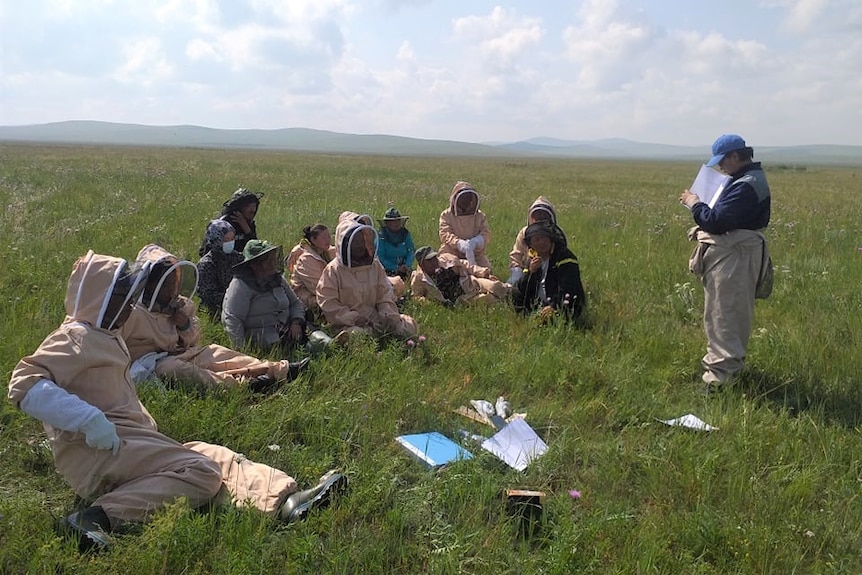 A woman in beekeeping gear talks to other people sitting in beekeeping gear on a vast plain.