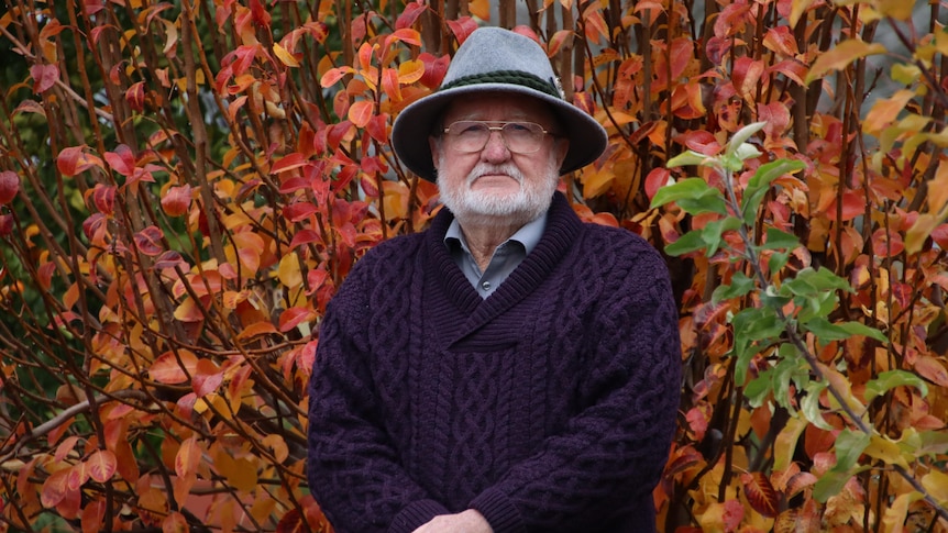 An older bearded man wearing a hat, glasses, shirt and jumper standing in front of a tree with multicoloured leaves