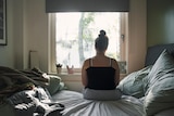 Seen from behind, a young person with singlet and hair tied in bun sits on a bed in slightly dark room facing a large window.