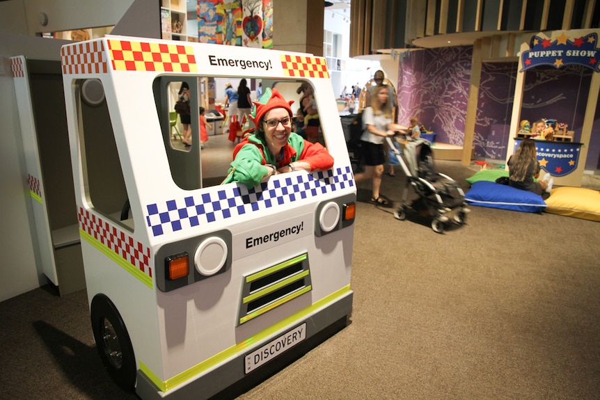 A woman wearing an elf outfit smiles from a play designed emergency vehicle