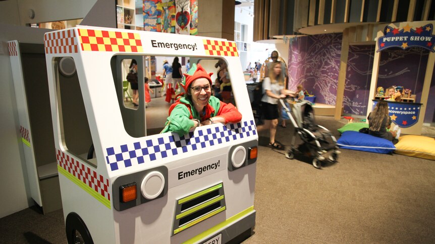 A woman wearing an elf outfit smiles from a play designed emergency vehicle