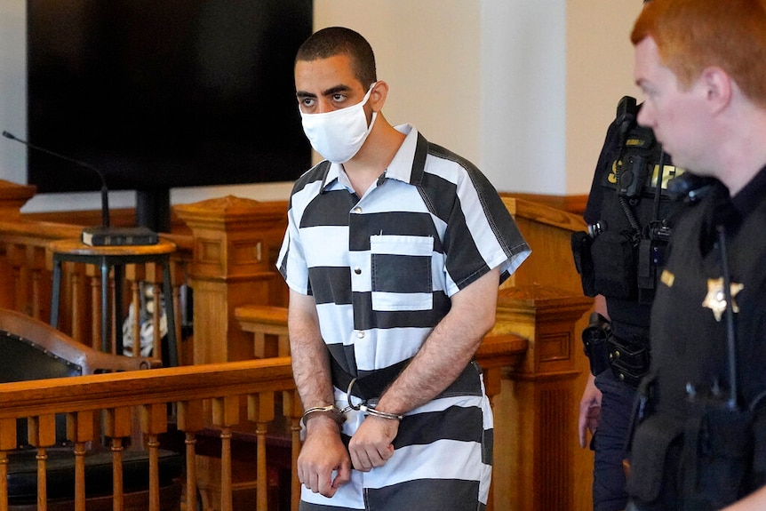 Hadi Matar, 24, center, arrives for an arraignment in cuffs, wearing black and white prison clothes and a mask.