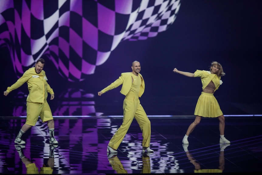 Three people dance on a purple stage with a checkerboard behind them. They're all wearing yellow.