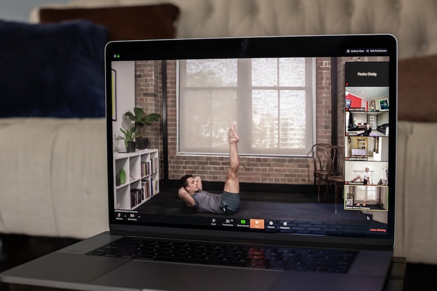 A laptop with a zoom dance class, an instructor on the floor doing a stretch as others join in