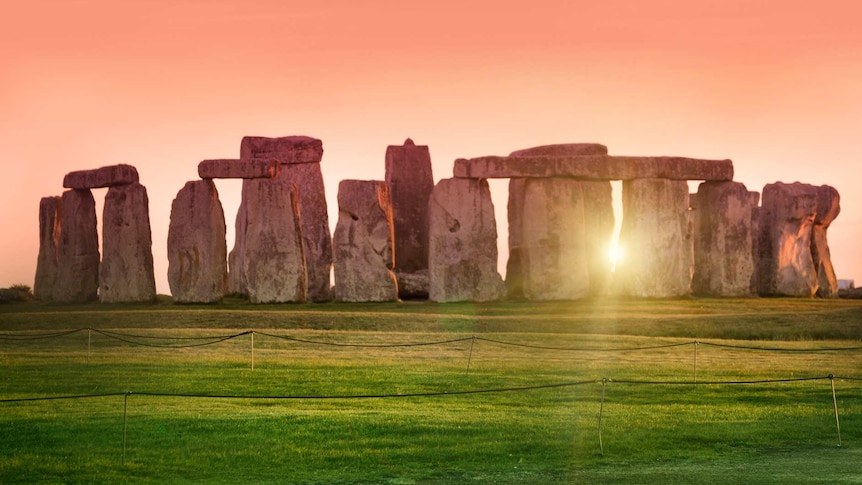 Photo of Stonehenge prehistoric monument at sunset. Focus on grass in forefront, stone monument slightly blurred in background.