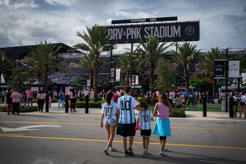 A family wearing blue and white striped Messi jerseys cross the road towards DRV PNK Stadium sign and crowds