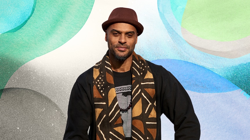 MC N'fa Jones stands in front of a colourful illustrated background wearing a hat and scarf