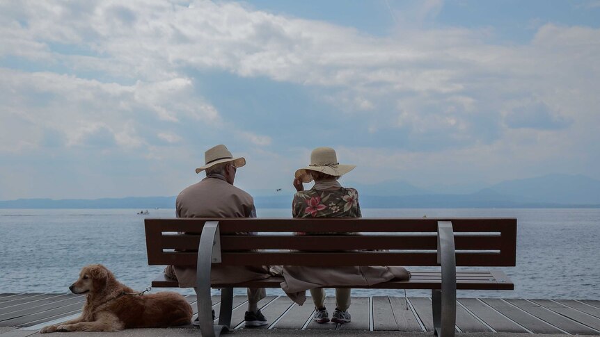 Two elderly people sit side by side on a bench overlooking a harbour, the man holding the leash of a Labrador.