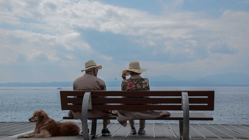 Two older people sit on a bench with their dog, looking out at the ocean
