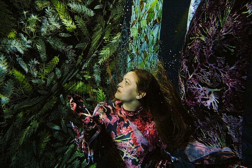 A woman in a bodysuit printed with pink seaweed swims under water among printed silk fabrics