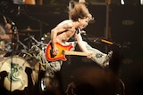 A long-haired man jumps while playing a yellow and red guitar.