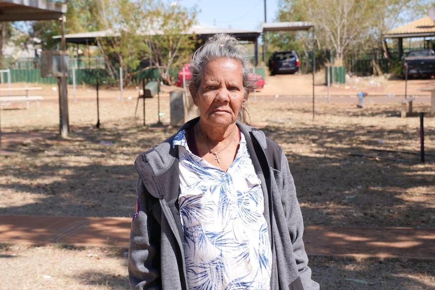 An older Indigenous woman wsearing a white and blue shirt and grey cardigan stands in a park.