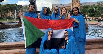 Seven women dressed in blue pose with a Sudanese flag in front of a fountain and cathedral in Sydney,