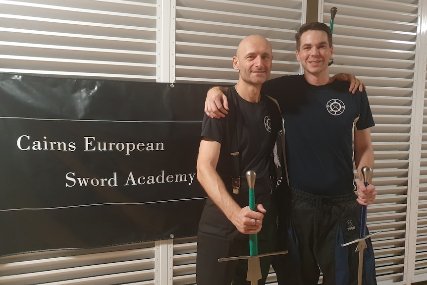 Two men stand next to a sign holding swords