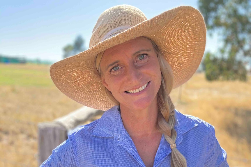 A woman with a blonde plait, wearing jeans, a blue shirt and a wide-brimmed hat, stands in front of a fence and smiles.