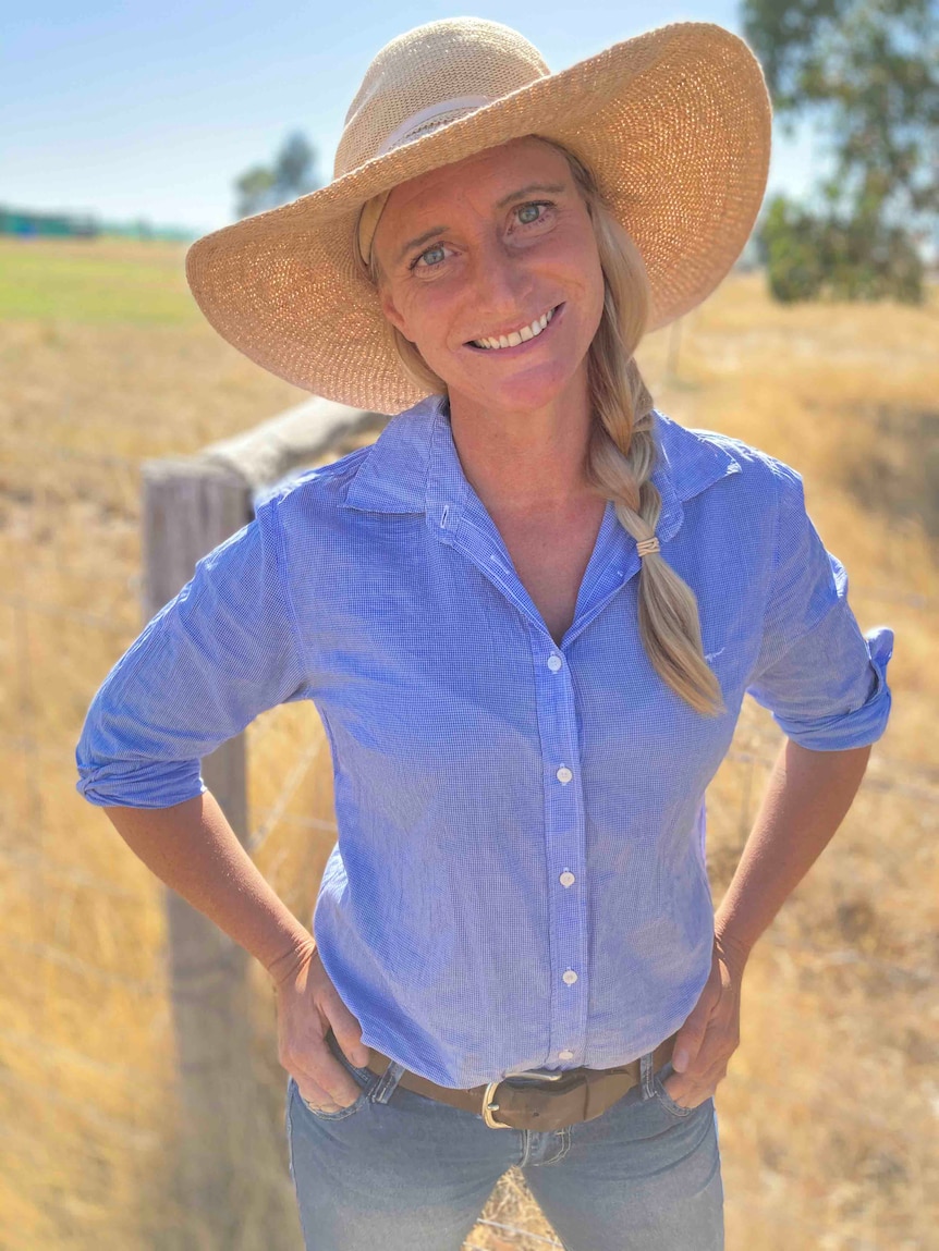 A woman with a blonde plait, wearing jeans, a blue shirt and a wide-brimmed hat, stands in front of a fence and smiles.