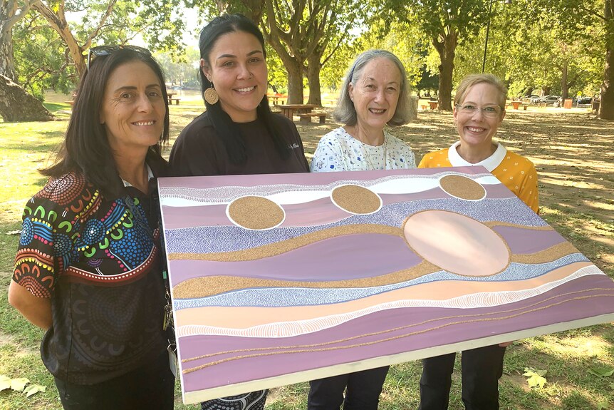 Megan Clayton,  Teisha Maksymow-McGuiness, Diane Davey and Kerry Strauch hold up Teisha's artwork in a parklands.
