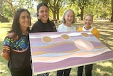 Megan Clayton,  Teisha Maksymow-McGuiness, Diane Davey and Kerry Strauch hold up Teisha's artwork in a parklands.