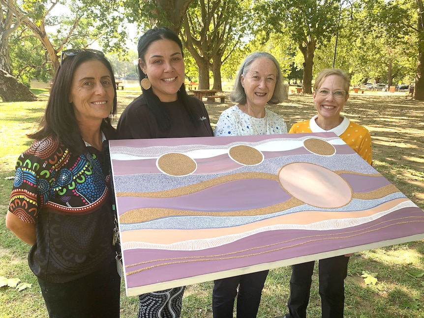 Megan Clayton,  Teisha Maksymow-McGuiness, Diane Davey and Kerry Strauch hold up Teisha's artwork in a parklands.