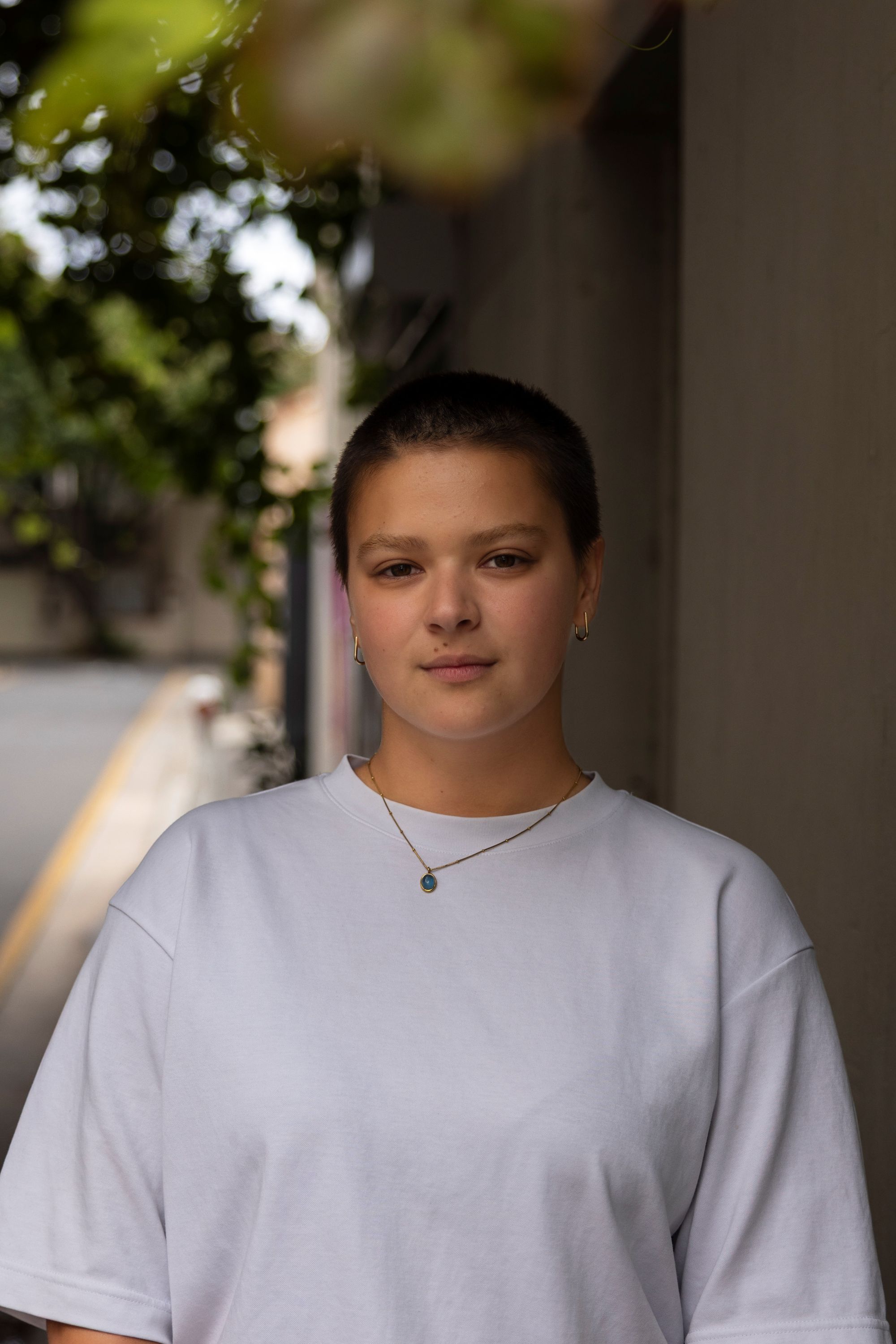 A nonbinary person with a shaved head, wearing a necklace and a white t-shirt.