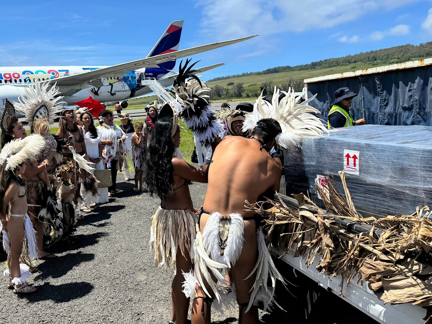 People of Rapa Nui dressed in traditional clothing welcome skeleton remains home in black boxes, plane in background. 