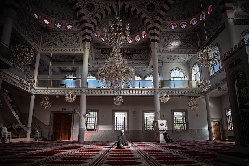 Imam Osman Boyaci sits on red patterned carpet inside Auburn Gallipoli Mosque during daytime.