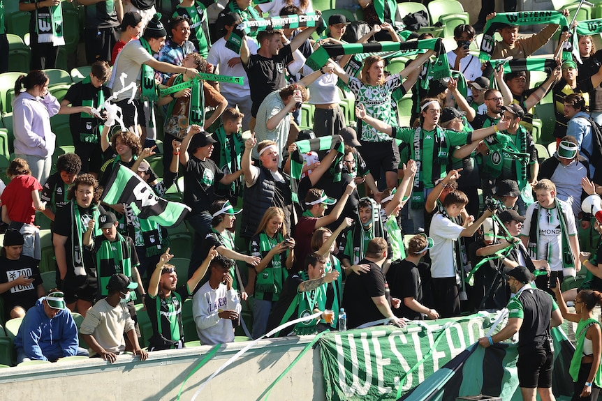 Soccer fans wearing green and black hold up scarves in the stands