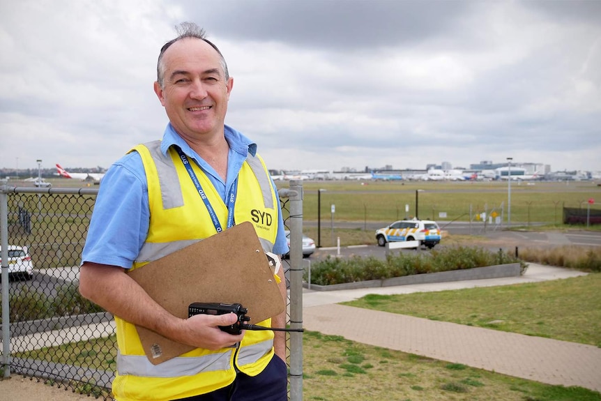 Nigel Coghlan at the Shep's Mound plane spotting spot site at Sydney Airport holding a clipboard with planes behind him.