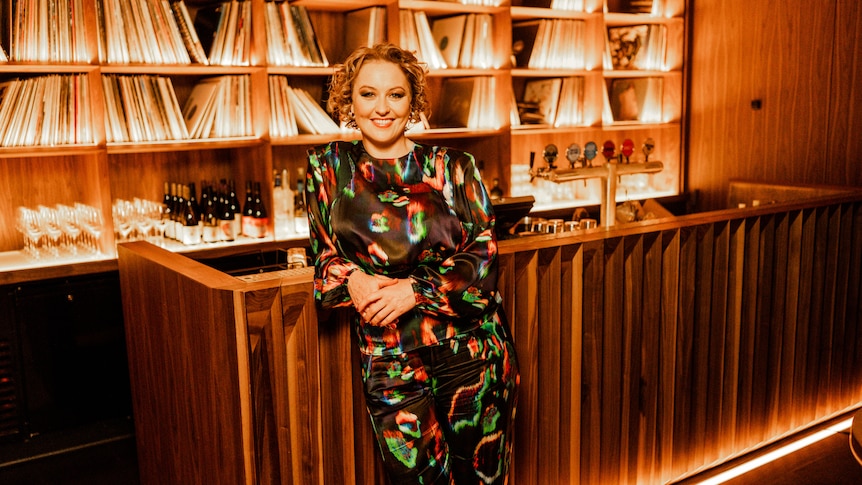 Zan Rowe leans against a wooden bar wearing a colourful dress. A record collection and bottles of wine are in the background.