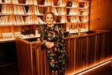 Zan Rowe leans against a wooden bar wearing a colourful dress. A record collection and bottles of wine are in the background.