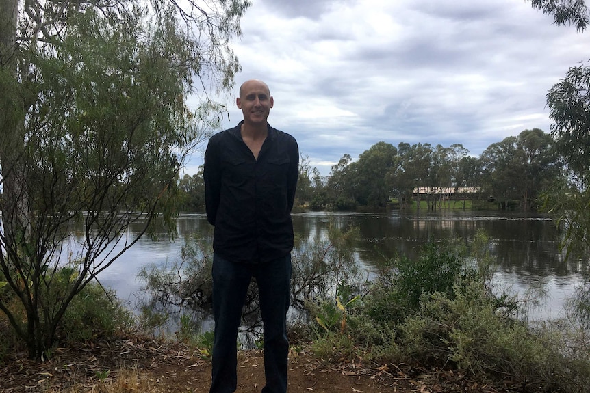 A bald man, wearing black shirt and trousers, hands behind back, stands by a river, vegetation around him, blue, cloudy sky.