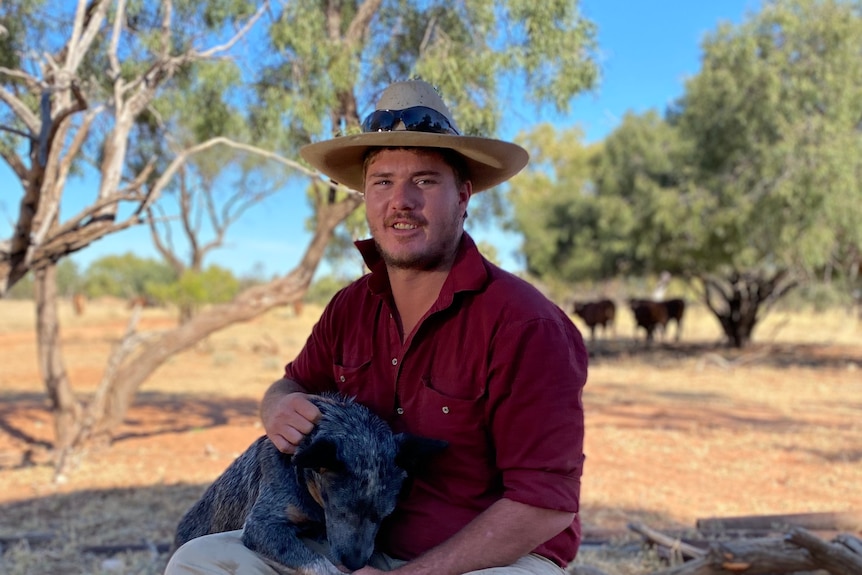 A man on a farm holding a dog, with cows in the background.