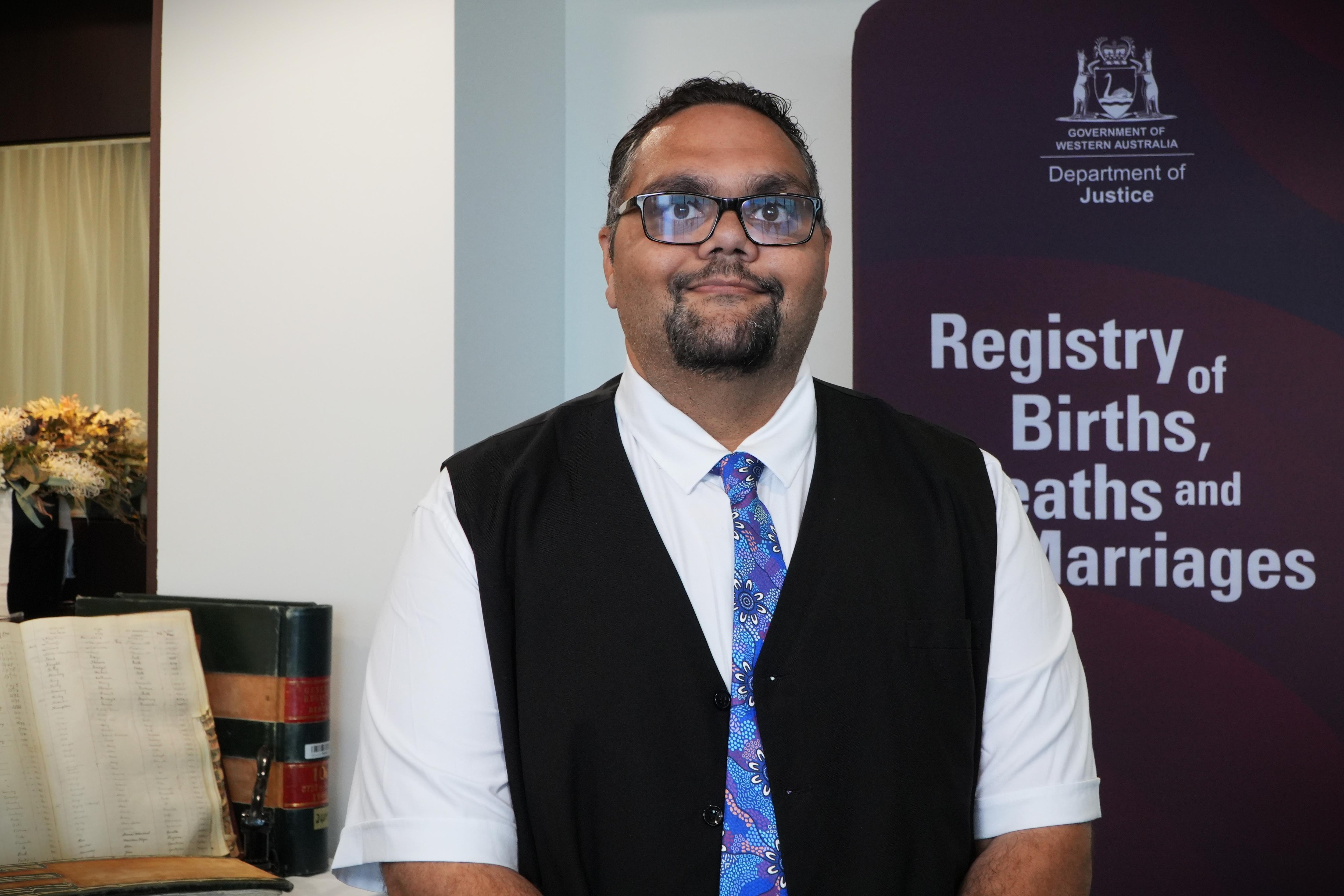 Un hombre con chaleco negro, camisa blanca y corbata sonríe a la cámara, detrás de él hay un cartel que dice 