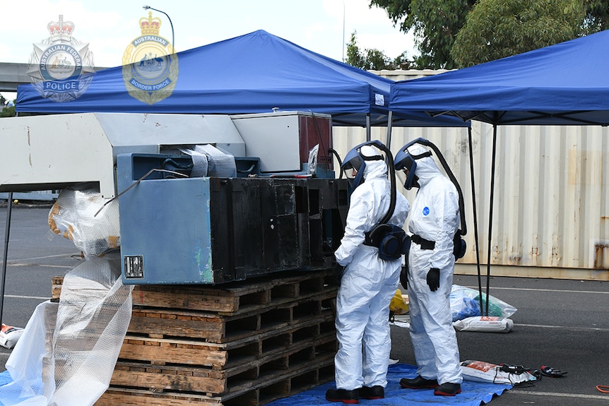 Two officers wear full PPE and masks as they look at industrial machinery.