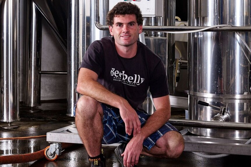 Man, centre of photo, kneels down in brewhouse with fermenters behind him in a sustainable brewery in Victoria