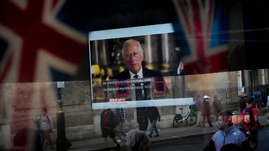 King Charles III's speech is seen on a television screen through a pub window reflecting Union Jacks and people on a street
